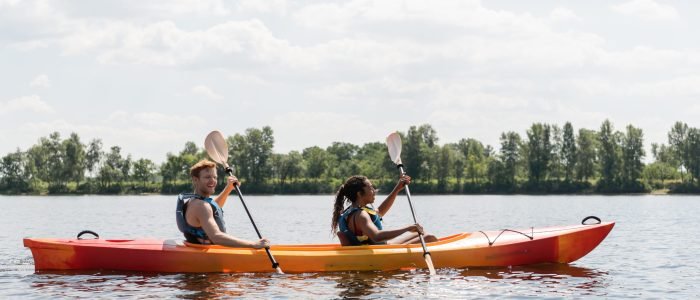 side view of smiling redhead man and brunette african american woman in life vests spending summer weekend by sailing in kayak on river under cloudy sky in summer